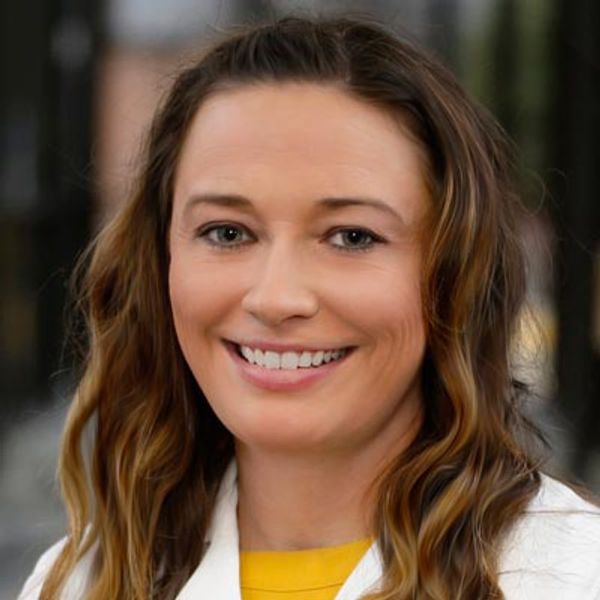 Smiling woman with long wavy hair, wearing a white coat and yellow top.