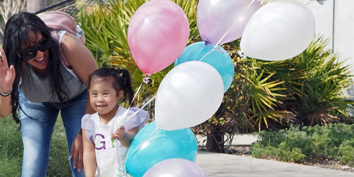 Smiling child holding colorful balloons with a woman waving beside her outdoors.