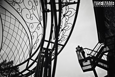 A couple shares a kiss on a fire escape against a cloudy sky, with intricate wrought iron designs in the foreground.