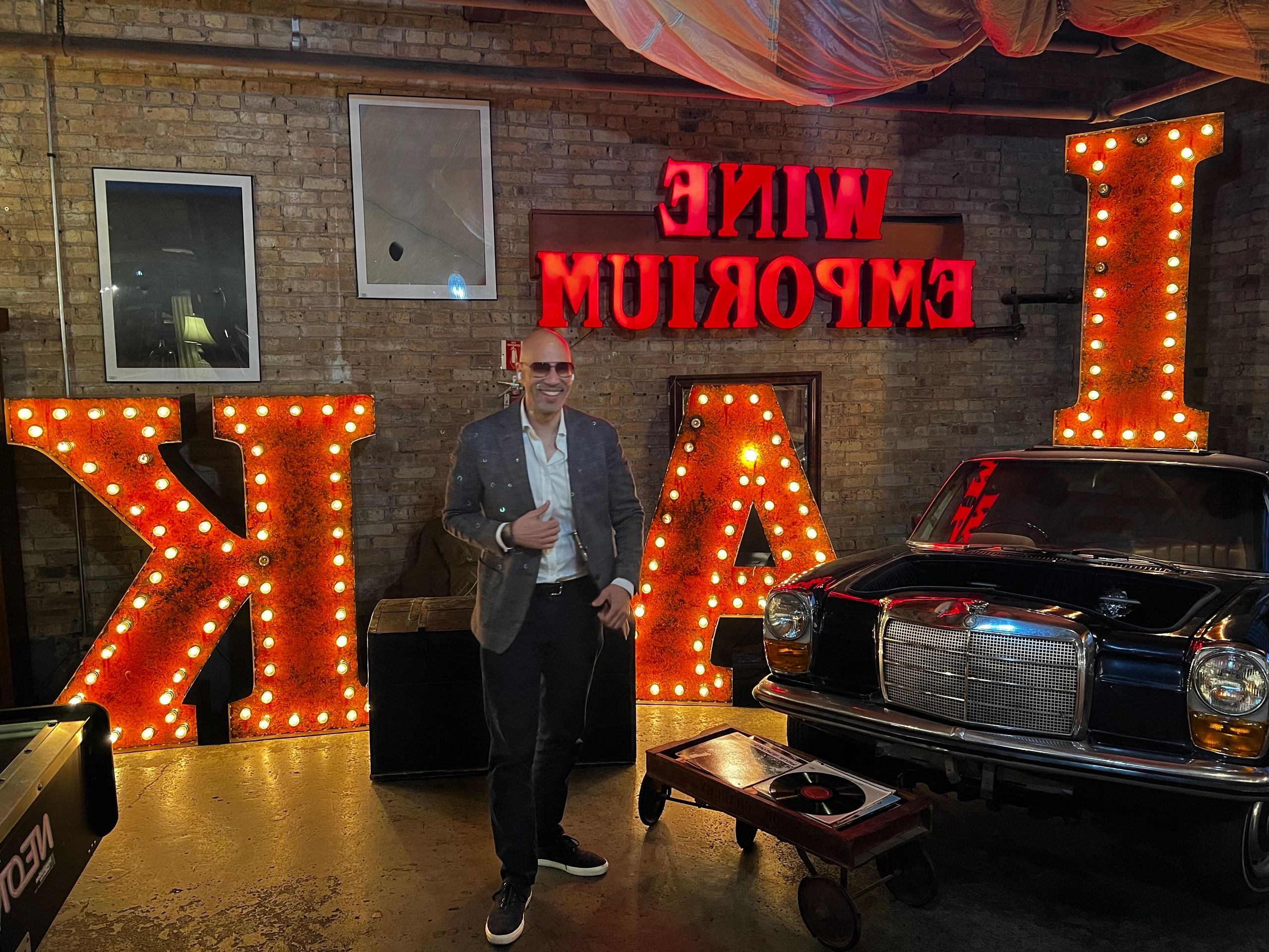 Man posing in front of illuminated letters and a vintage car in a brick-walled room.