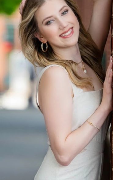 A young woman with blonde hair smiles while leaning against a brick wall.