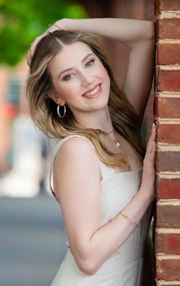 A young woman with blonde hair smiles while leaning against a brick wall.
