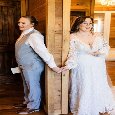 Two brides hold hands around a wooden corner, sharing a quiet moment before their wedding.