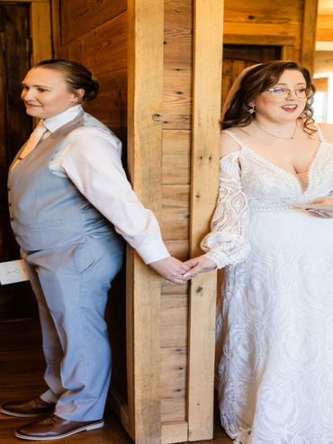 Two brides hold hands around a wooden corner, sharing a quiet moment before their wedding.