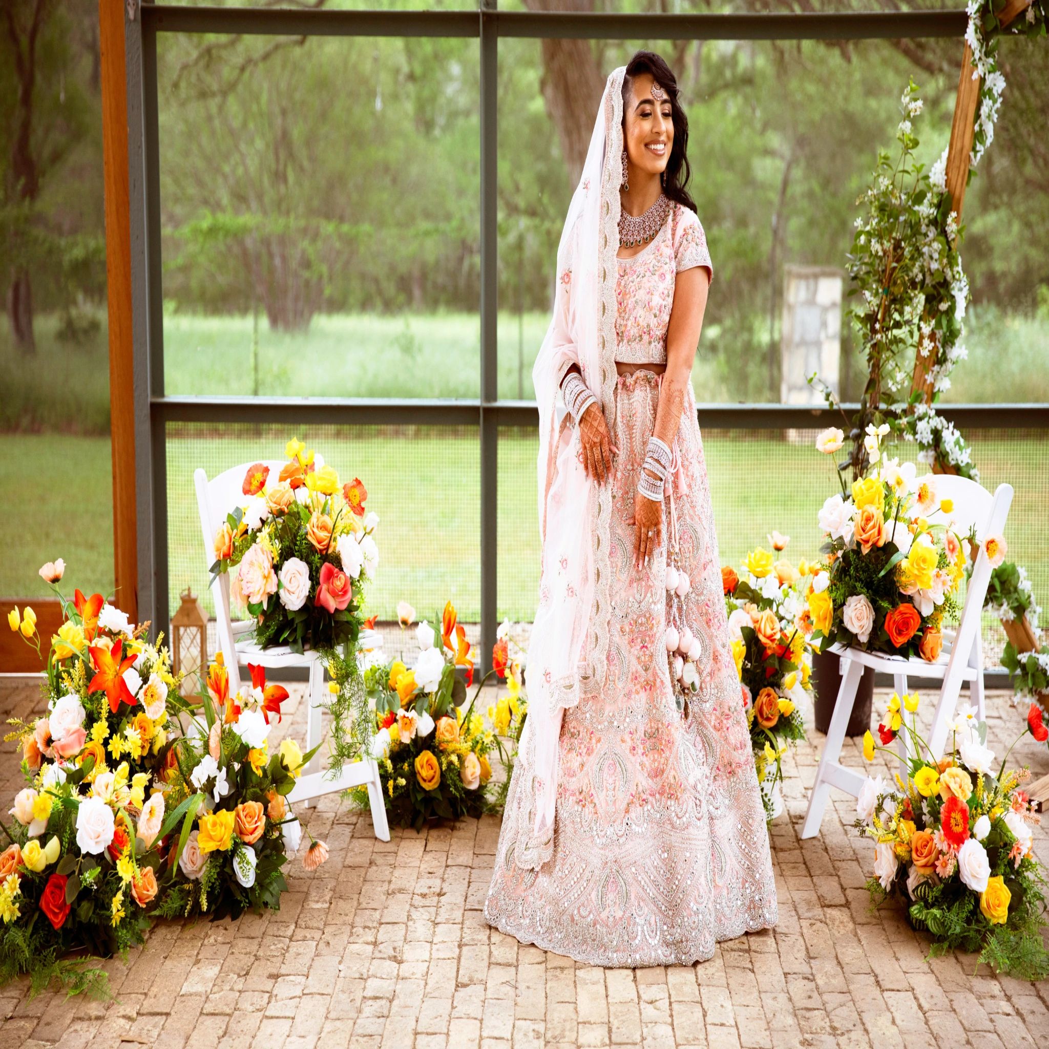 A bride in traditional attire stands smiling among floral decorations.