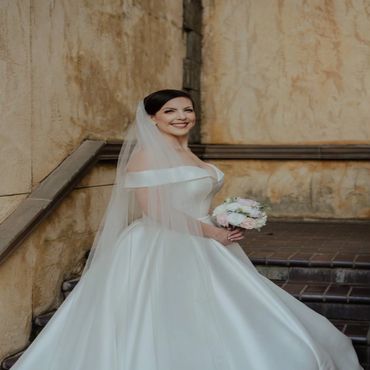 A joyful bride in an elegant white gown holds a bouquet, smiling against a rustic wall.