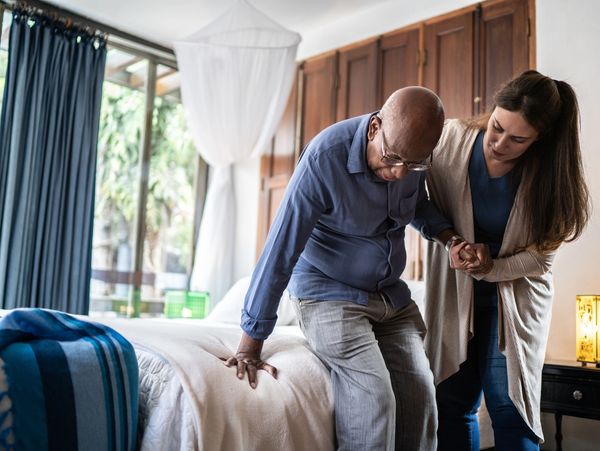 A young woman wearing scrubs helping a senior man sit or stand near a bed.