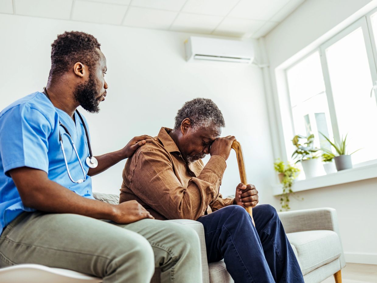 Young man wears scrubs and a stethoscope comforts a senior man holding a cane, they sit on a couch.