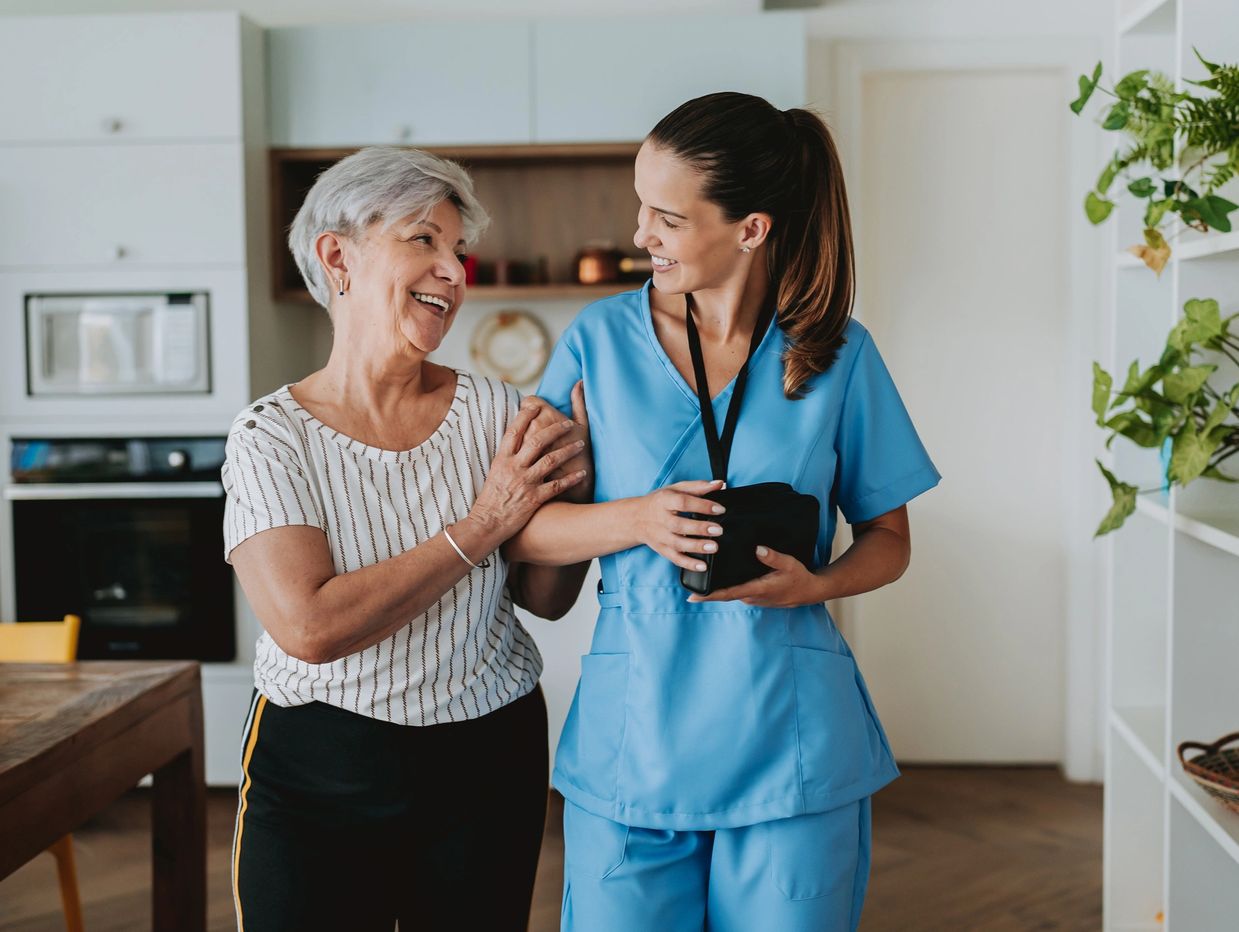 A senior woman with short white hair embraces the arm of a young woman wearing scrubs in a kitchen.