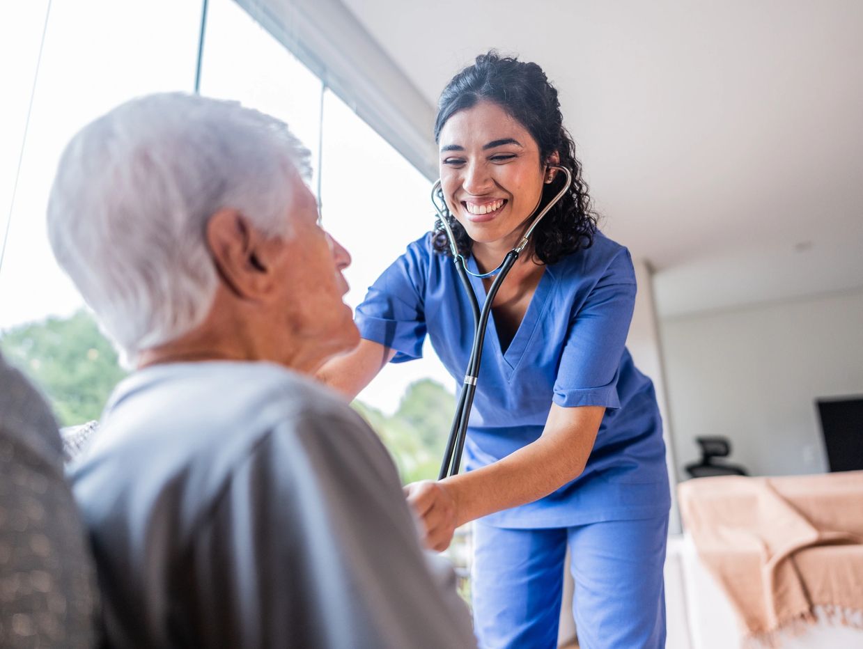 A young woman wearing scrubs uses a stethoscope on an older man seated in a well lit room.