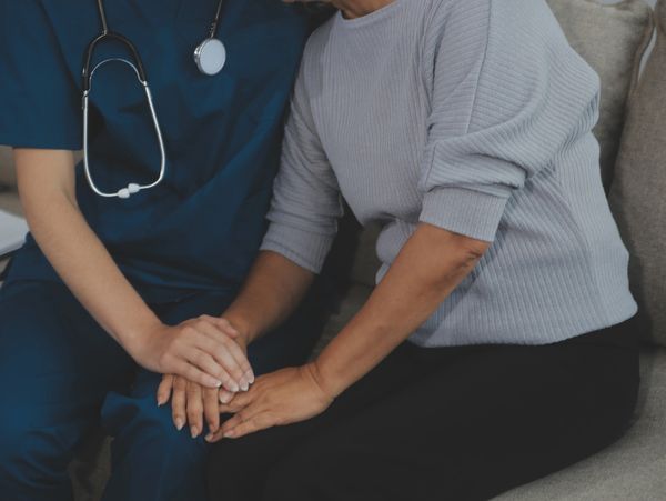 A young person wearing scrubs and a stethoscope embracing an older woman as they sit on a couch.