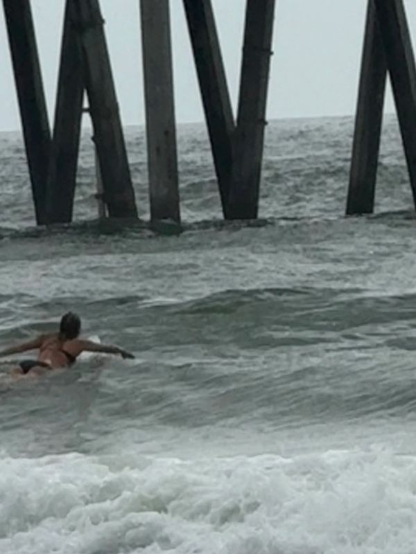 Person paddling on a surfboard near a pier in choppy ocean waters.