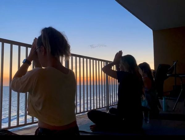 Three people practicing yoga on a balcony at sunset overlooking the ocean.