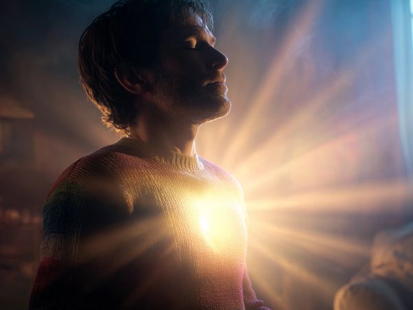 Man meditates with glowing light radiating from his chest in a serene room.