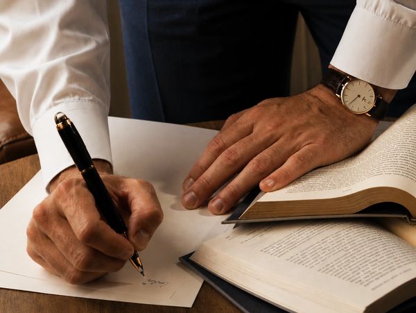 A person writing with a pen beside open books on a wooden table.