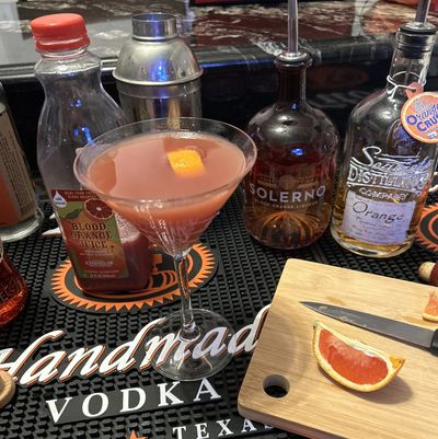 A pink cocktail with an orange peel garnish on a bar mat surrounded by cocktail ingredients.