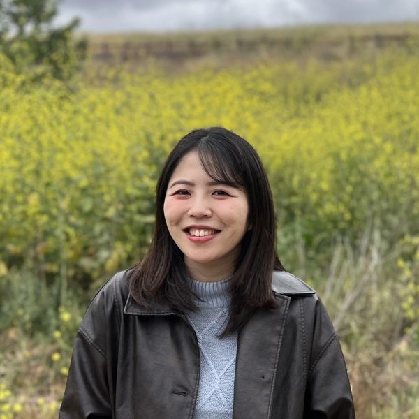 A smiling woman in a leather jacket stands in front of yellow flowers.