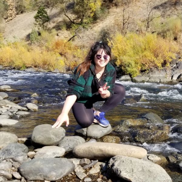 Young woman crouching on rocks in a river, smiling and making a peace sign.
