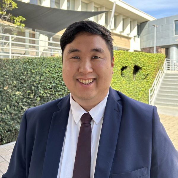 A smiling man in a suit and tie outdoors in a sunny urban setting.