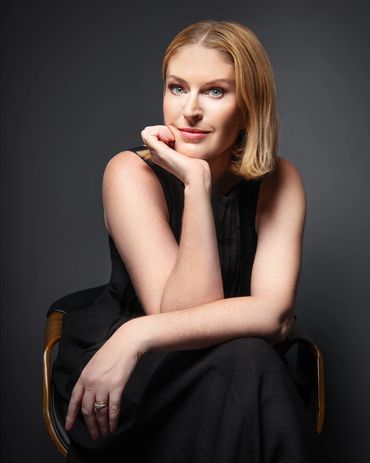 Studio editorial portrait of a blonde haired business woman wearing a black dress sat on a black lea