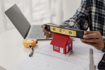 Person measures a red model house with a level tool on blueprints.