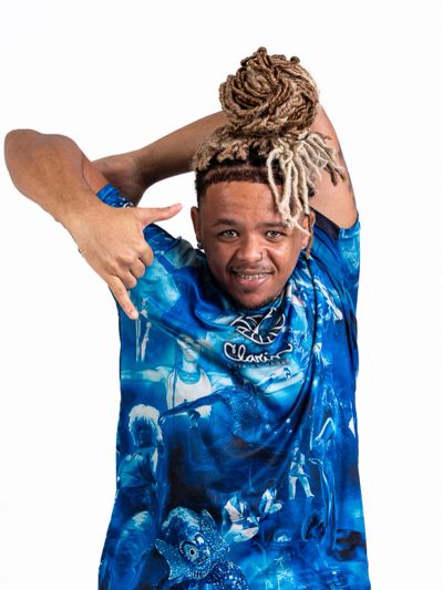 Man with unique dreadlocks in a blue graphic t-shirt making a hand gesture.