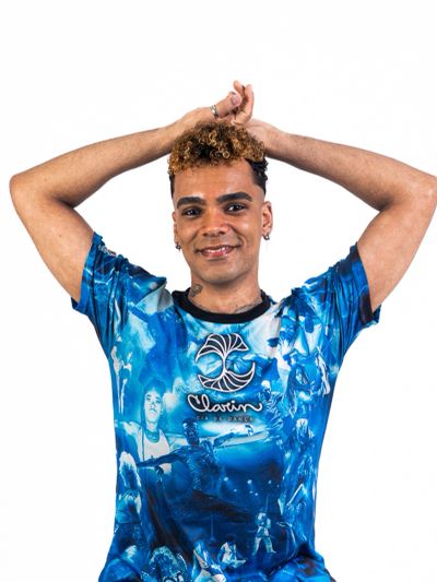 Young man with curly hair wearing a blue graphic t-shirt, smiling with hands raised behind his head.