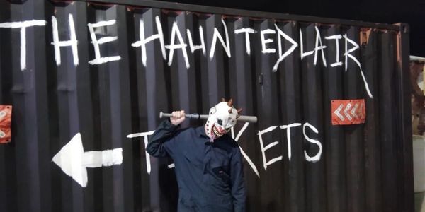 A person in a scary mask stands in front of a sign for The Haunted Lair tickets.