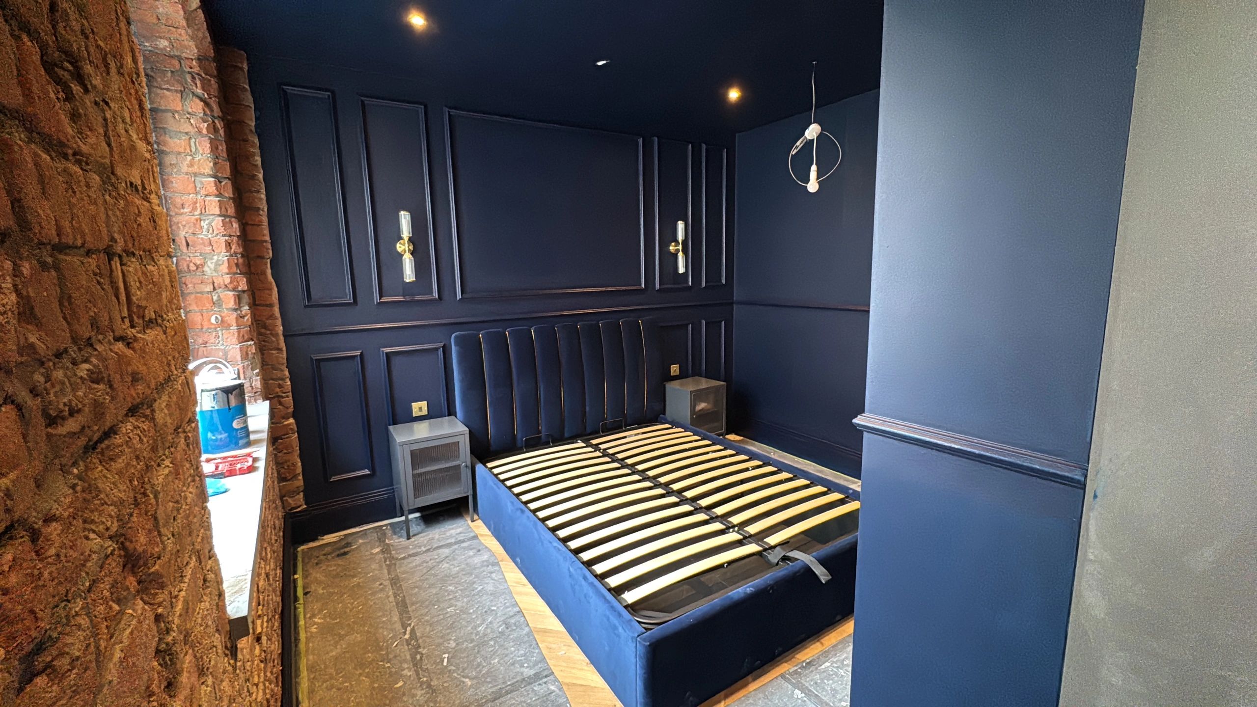 A dark blue bedroom under renovation with exposed brick and a bed frame without a mattress.