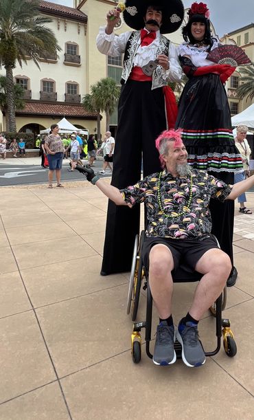 Randy in his wheelchair with stilt walkers beside him.