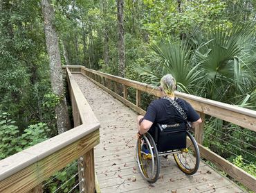 Randy in his wheelchair showing accessibility on a nature trail boardwalk. He is surrounded by trees.