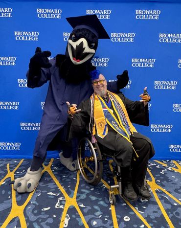 Randy in a wheelchair at his graduation giving a thumbs up as Sammy Seahawk poses behind him.