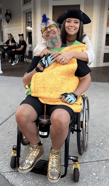 Randy in his wheelchair is dressed up as a taco and his wife is a pirate. She is hugging his neck.
