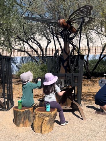 Two children wearing hats play with a mechanical insect sculpture outdoors.
