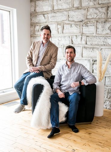 Two guys sitting on a chair in front of a grey brick wall.