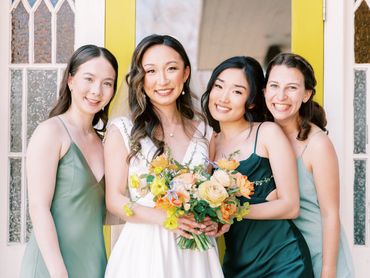 Bridesmaids in green, and bride posing for a photo in front of a yellow door holding wildflowers.