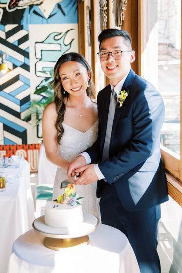Newlyweds posing for a photo in front of a graffiti wall while they cut their single layer cake.