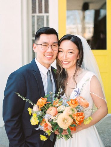 Newlyweds posing in front of a yellow door holding a bouquet of wildflowers.