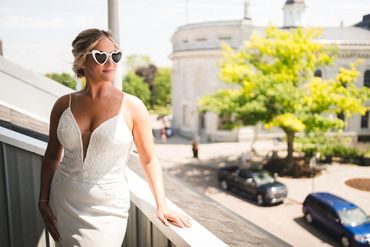 Bride wearing heart shaped glasses standing on a balcony takes in views of Kingston's Market Square