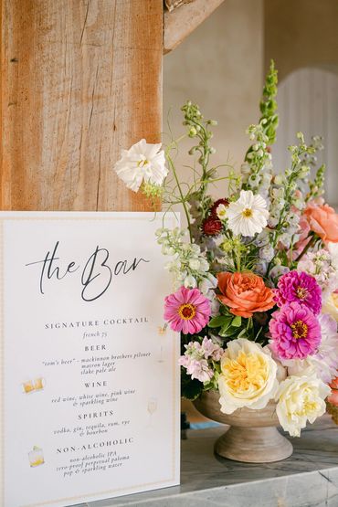 Welcome to the bar sign in white with black writing sitting next to a colorful summer flowers.