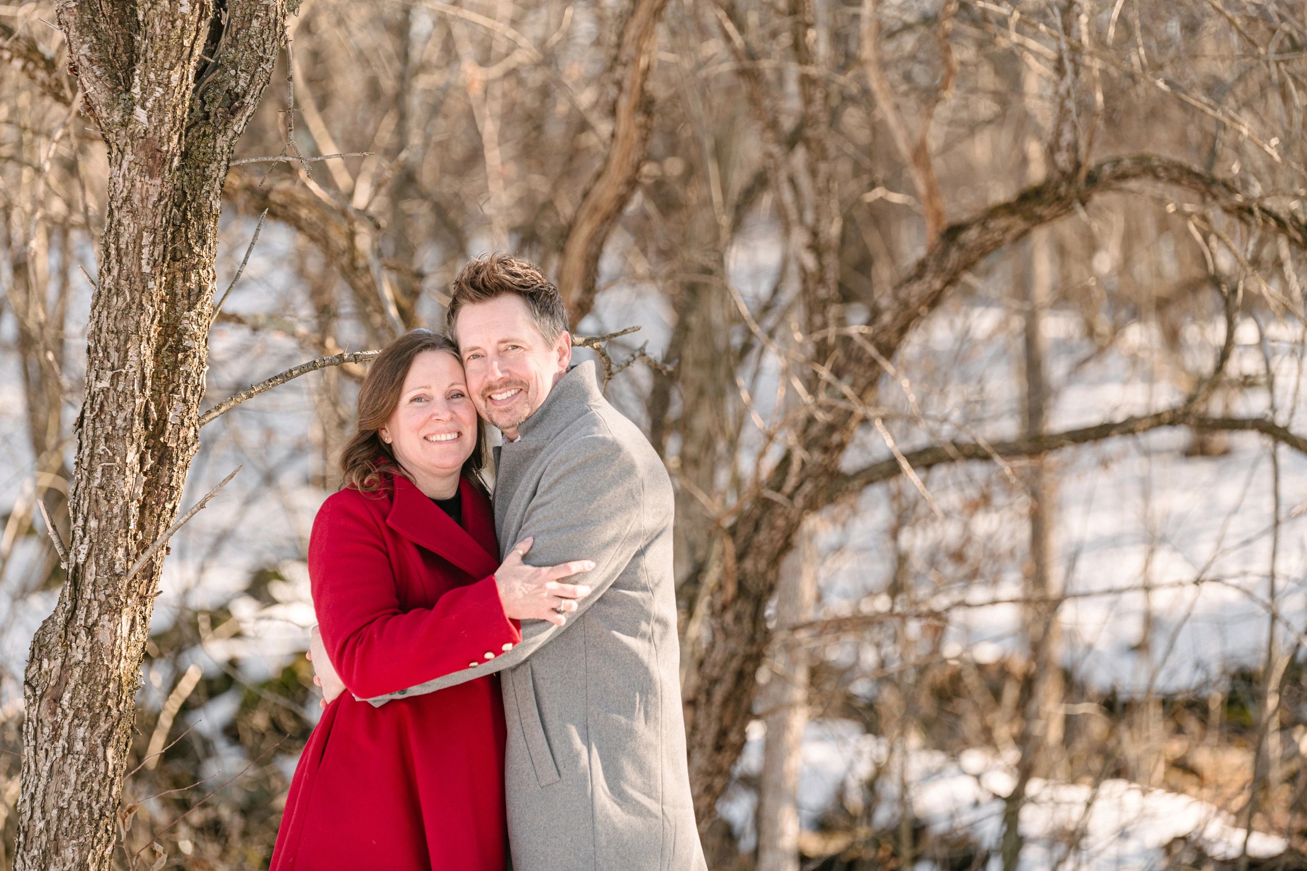 Girl wearing a red coat and a guy wearing a grey coat, hugging each other in a winter forest.