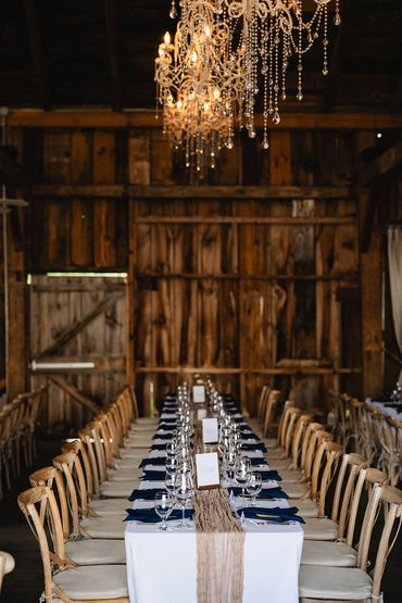 Chrystal chandaliers hang in a rustic barn on top of a perfectly set harvet table with blue napkins.