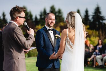 Officiant in brown suit speaking to a bride, groom and their guests.