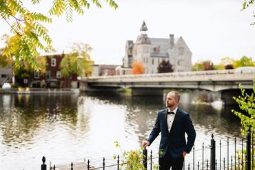 Groom standing an iron fence overlooking the river with the town of Almonte in the background