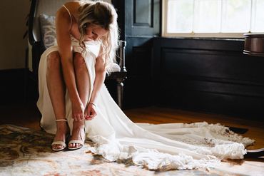 Bride does up here shoes while here dress drapes along the floor beside her.