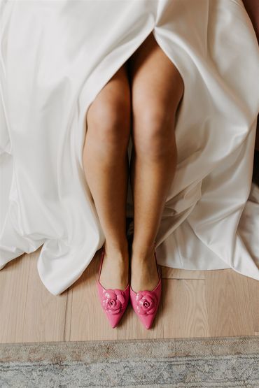 Brides legs sticking out of her white wedding dress wearing pink heal with rose details.