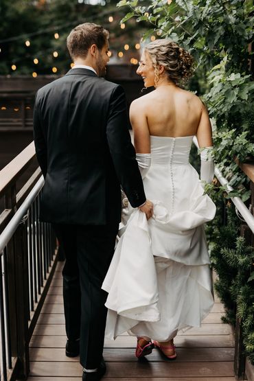 Groom holding brides dress while they walk up a ramp covered in vines.