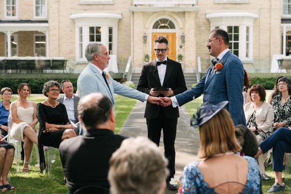 Grooms hold hands during a ceremony performed by an officiant dressed in a black tux.