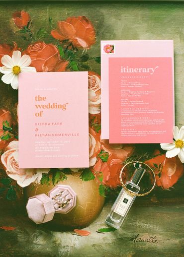 Pink inviations on a colorful floral backdrop plus their rings and perfume
