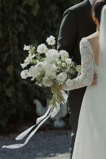 Bride in a lace dress, holding her white bouquet with the slik ribbon blowing in the wind.
